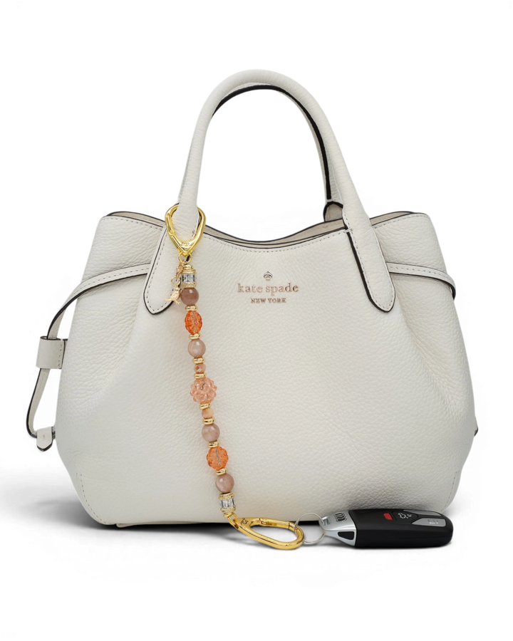 LD Keyfinder Birdsong Bliss beaded keychain with sunstone, coral flower beads, and gold bird charm on a white handbag.