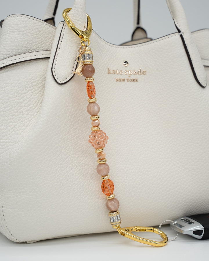 LD Keyfinder Birdsong Bliss beaded keychain with sunstone, coral flower beads, and gold bird charm on a white handbag.