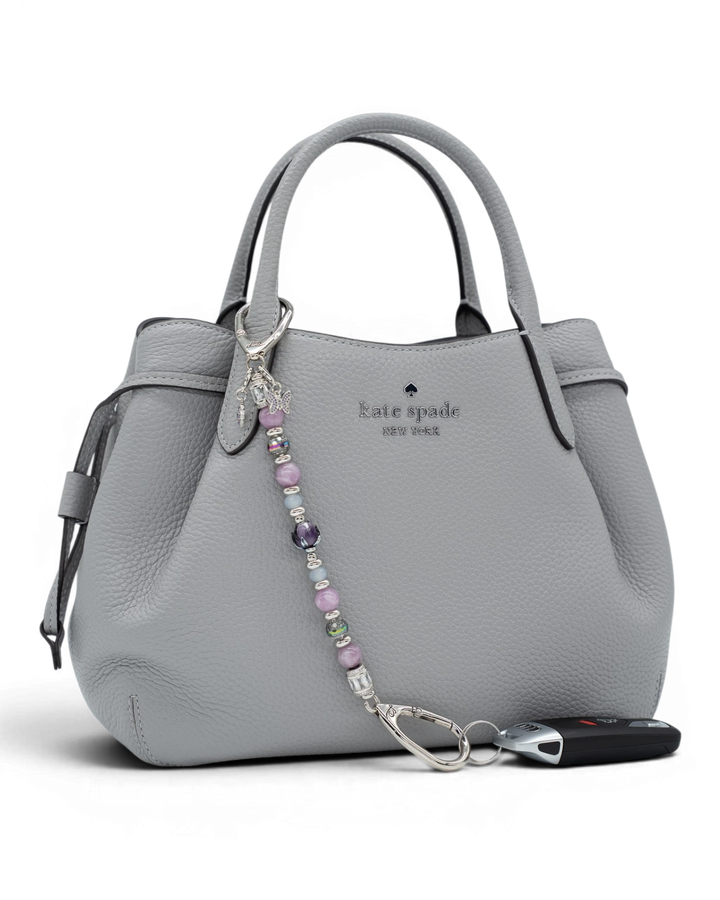 LD Keyfinder Blooming Joy beaded accessory with lavender lepidolite and glass charms attached to a grey leather handbag