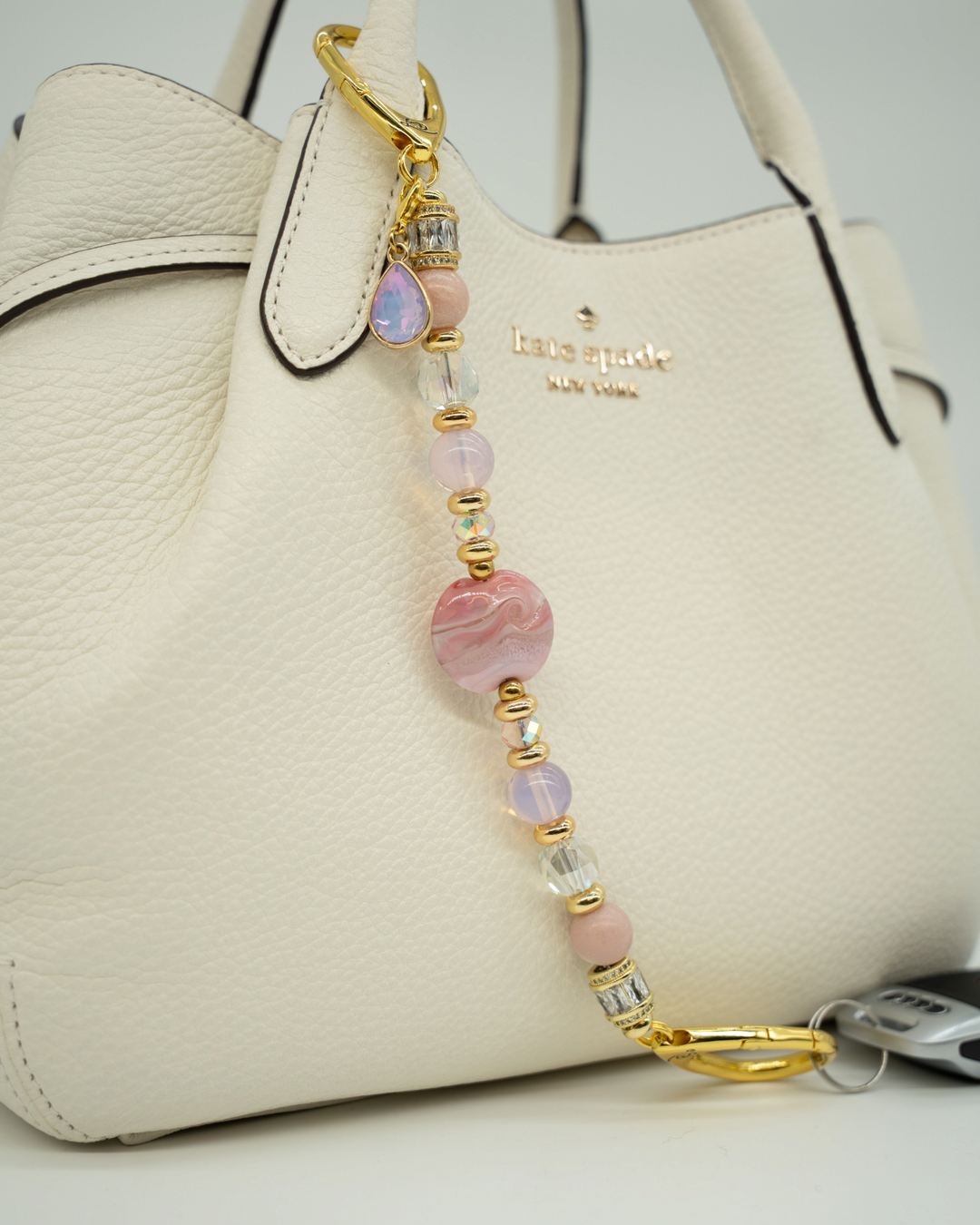 LD Keyfinder Blushing Opaline accessory with pink opal, artisan glass, and gold accents attached to a white handbag.