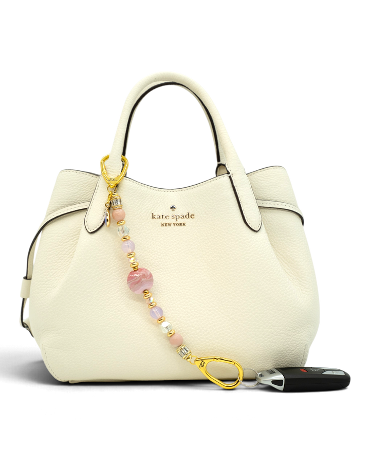 LD Keyfinder Blushing Opaline beaded accessory with pink opal and gold accents attached to a white leather handbag.