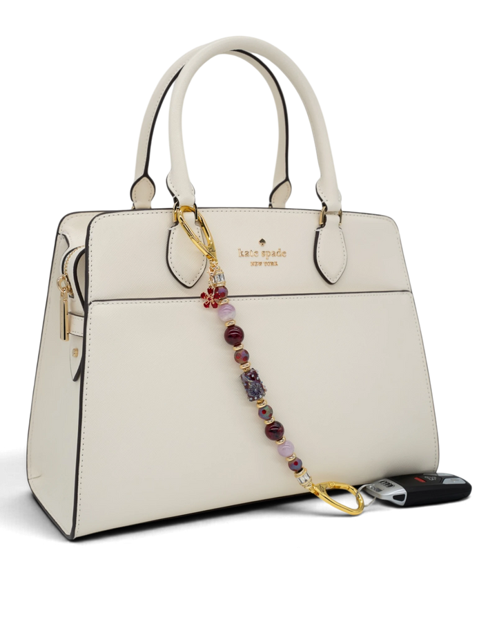 LD Keyfinder Bordeaux Bloom beaded accessory with burgundy agate, lavender kunzite, and gold flower charm on a handbag
