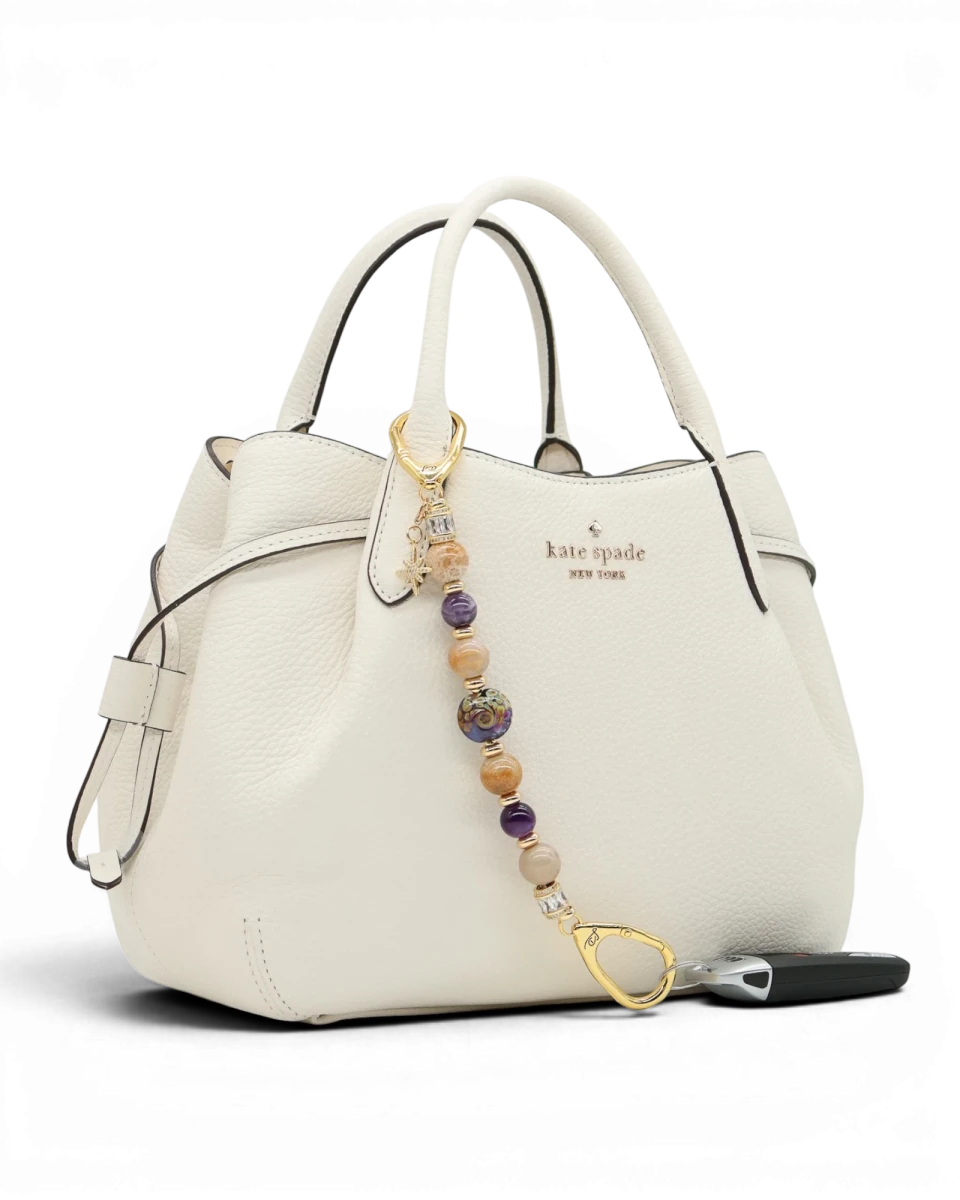 LD Keyfinder Golden Nebula charm with purple, gold, and amethyst beads, gold-plated star charm on white handbag.