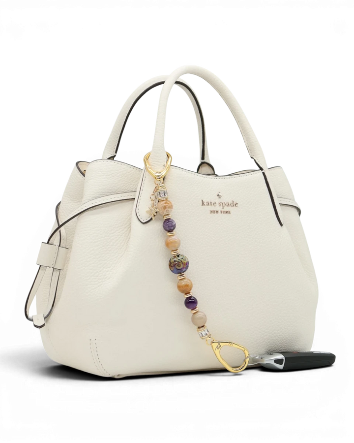 LD Keyfinder Golden Nebula charm with purple, gold, and amethyst beads, gold-plated star charm on white handbag.