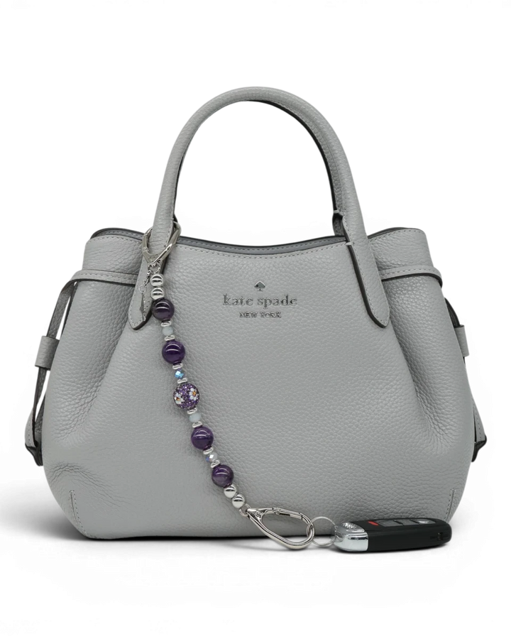 LD Keyfinder Hummingbird Dream charm with amethyst, jade glass beads, and hummingbird charm on a grey handbag.