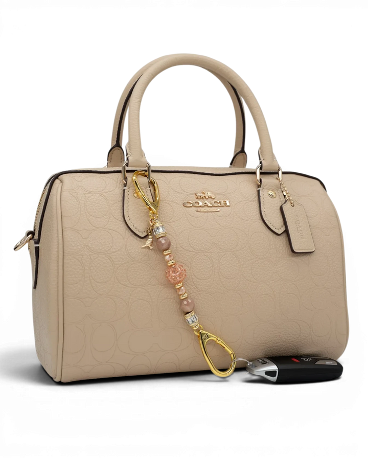 LD Keyfinder Petite Birdsong Bliss beaded charm with sunstone, peach glass, and gold bird accent on a beige handbag.