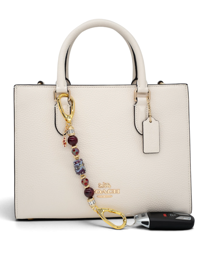 LD Keyfinder Petite Bordeaux Bloom accessory with burgundy agate, artisan glass, and gold charms on a white handbag.