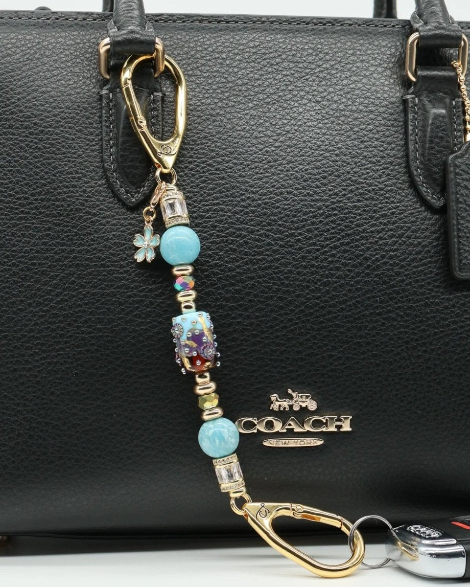 LD Keyfinder Petite Gilded Fantasy keychain with blue beads, gold accents, and a flower charm attached to a black handbag.