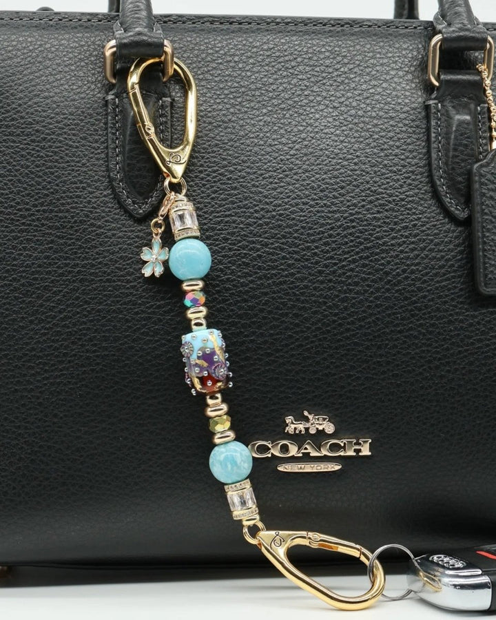 LD Keyfinder Petite Gilded Fantasy keychain with blue beads, gold accents, and a flower charm attached to a black handbag.