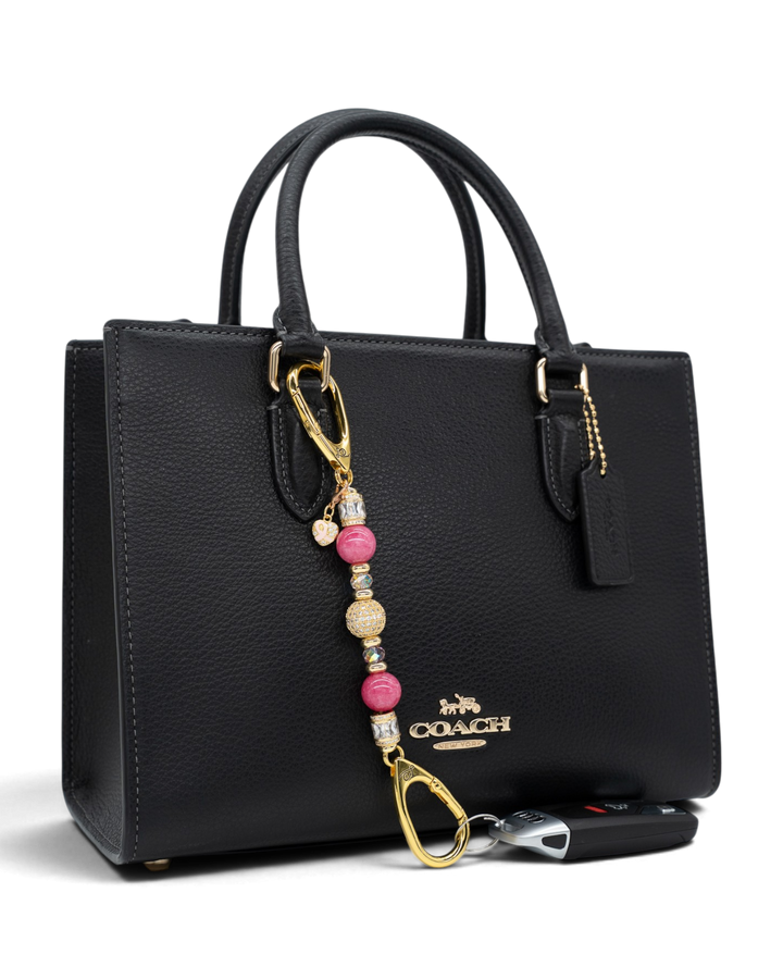 LD Keyfinder Petite Radiant Hope beaded charm with pink jade and gold accents attached to a black leather handbag.