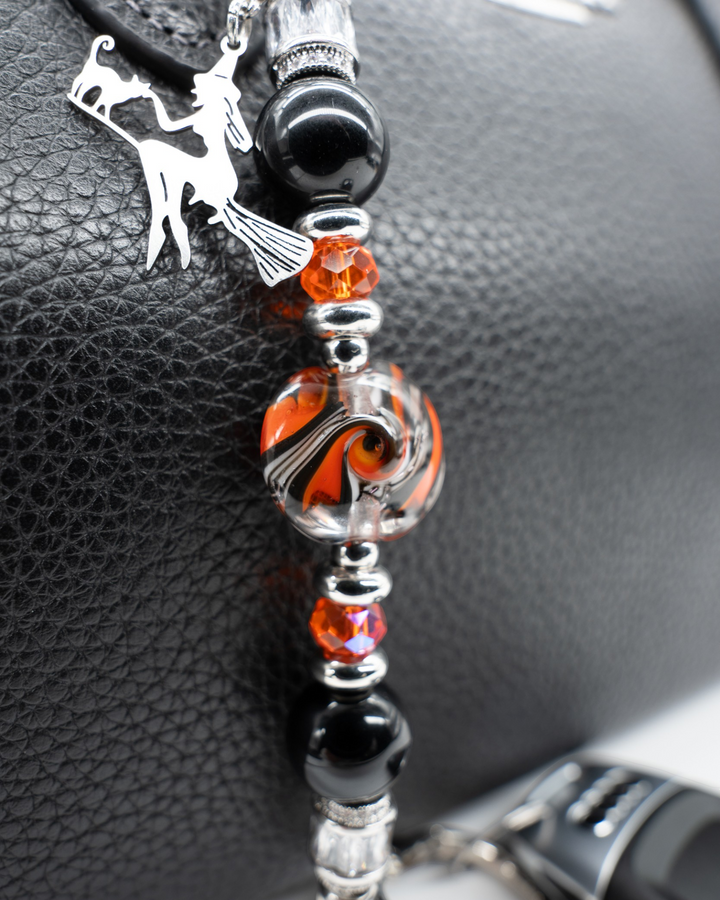 LD Keyfinder Petite Spellbound accessory with black obsidian, orange glass, and a witch charm on a black leather bag.